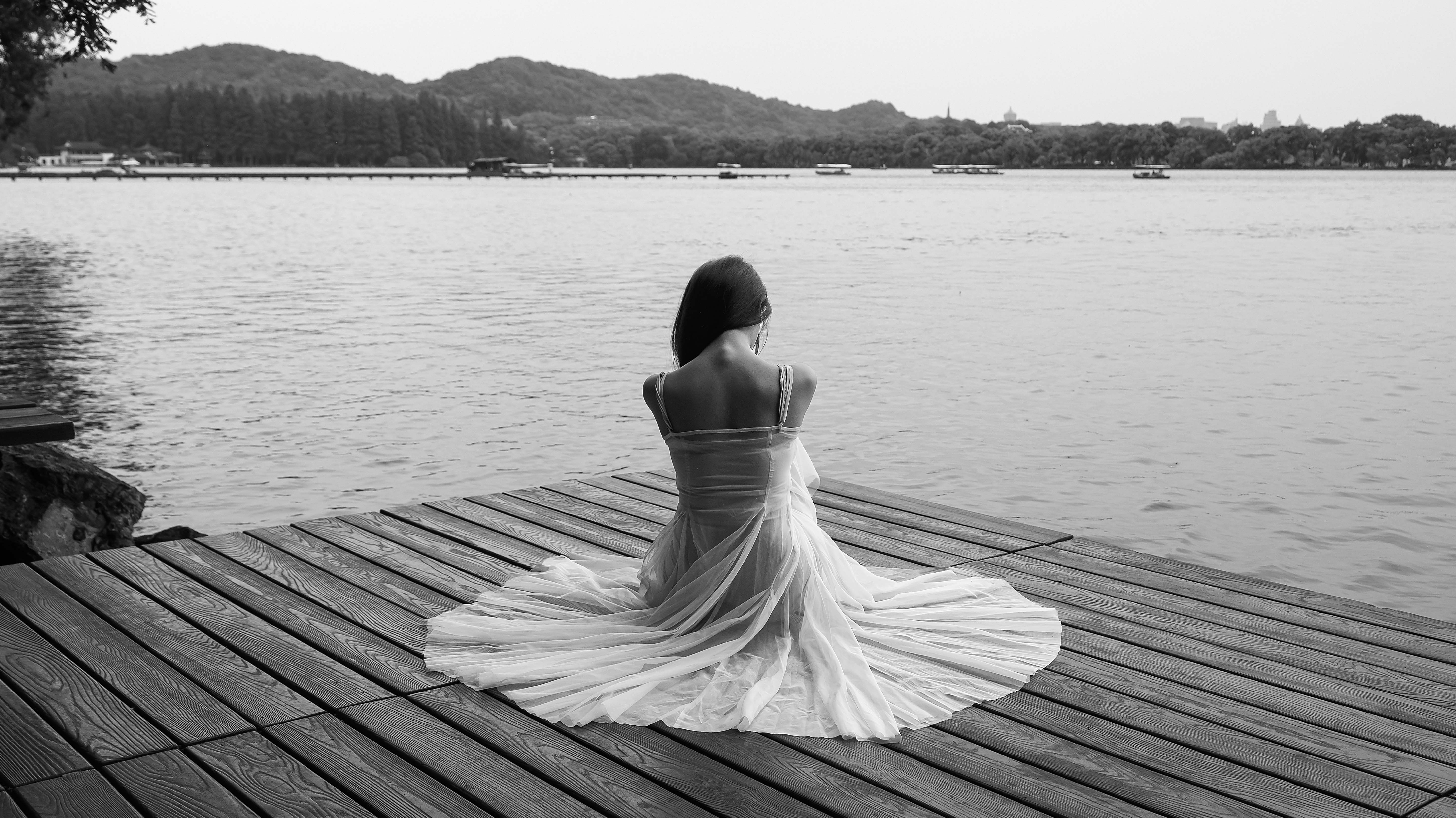 Woman in a flowing dress sits by a peaceful lake on a wooden pier in monochrome.