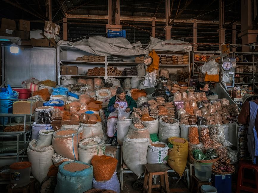 A Nigerian market stall displaying neatly arranged piles of ground egusi in open sacks, dried crayfish in a bowl, stockfish, and a yellow container of palm oil, with a cheerful market woman in a colourful ankara wrapper attending to a customer. Bright daylight, busy market atmosphere, Ibadan-style open-air market setting.