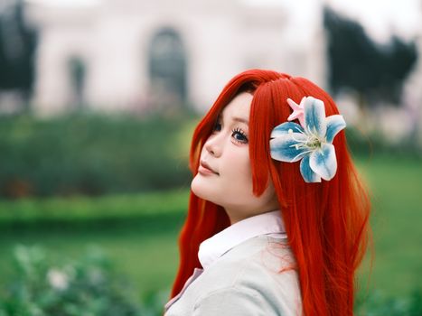 Colorful cosplay portrait of a woman with red hair and floral hair accessory in a serene park setting.