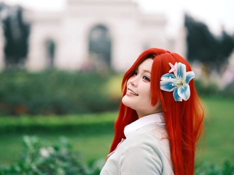 A smiling cosplayer with red hair and a floral accessory, captured in a lush outdoor setting.