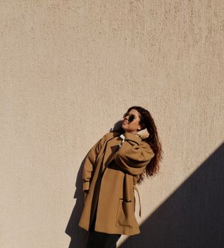 Elegant woman with curly hair and sunglasses standing in sunlight casting a shadow against a wall.