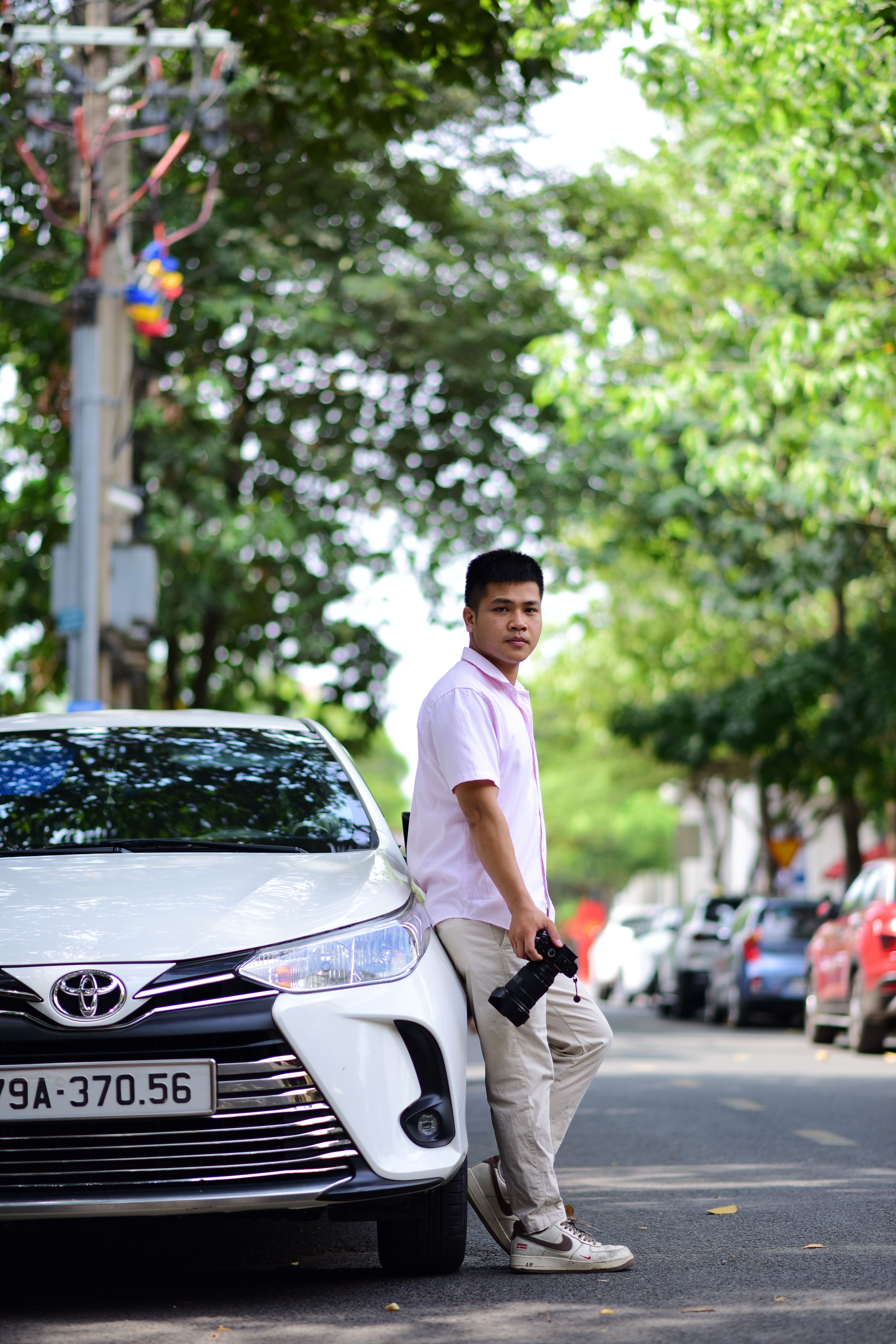 A young man stands casually with a camera, leaning on a white car in a leafy urban street.