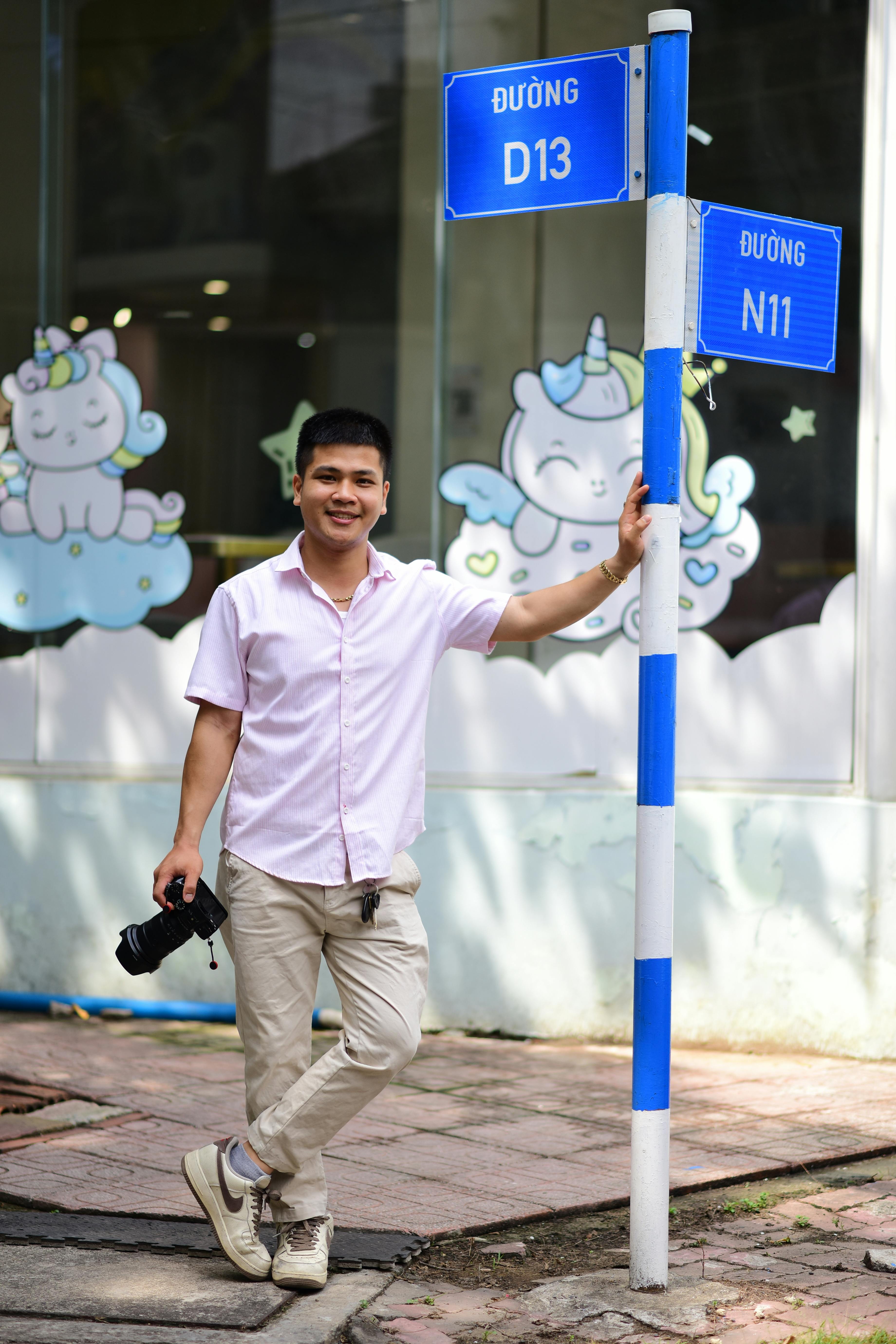 Photographer stands by a street sign on a sunny day in Vietnam, smiling.
