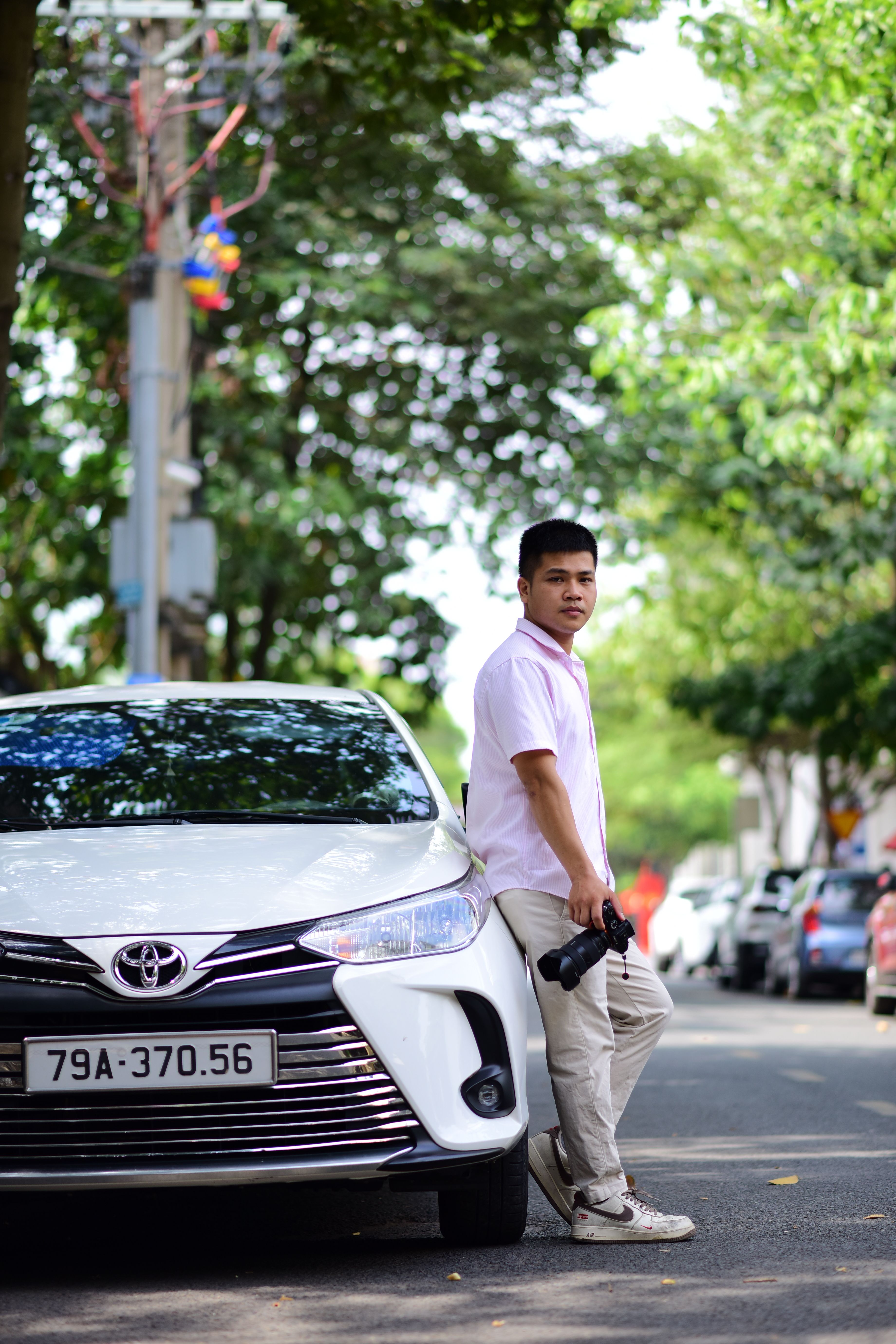 A young man in casual wear leaning against a car holding a camera outdoors on a sunny day.