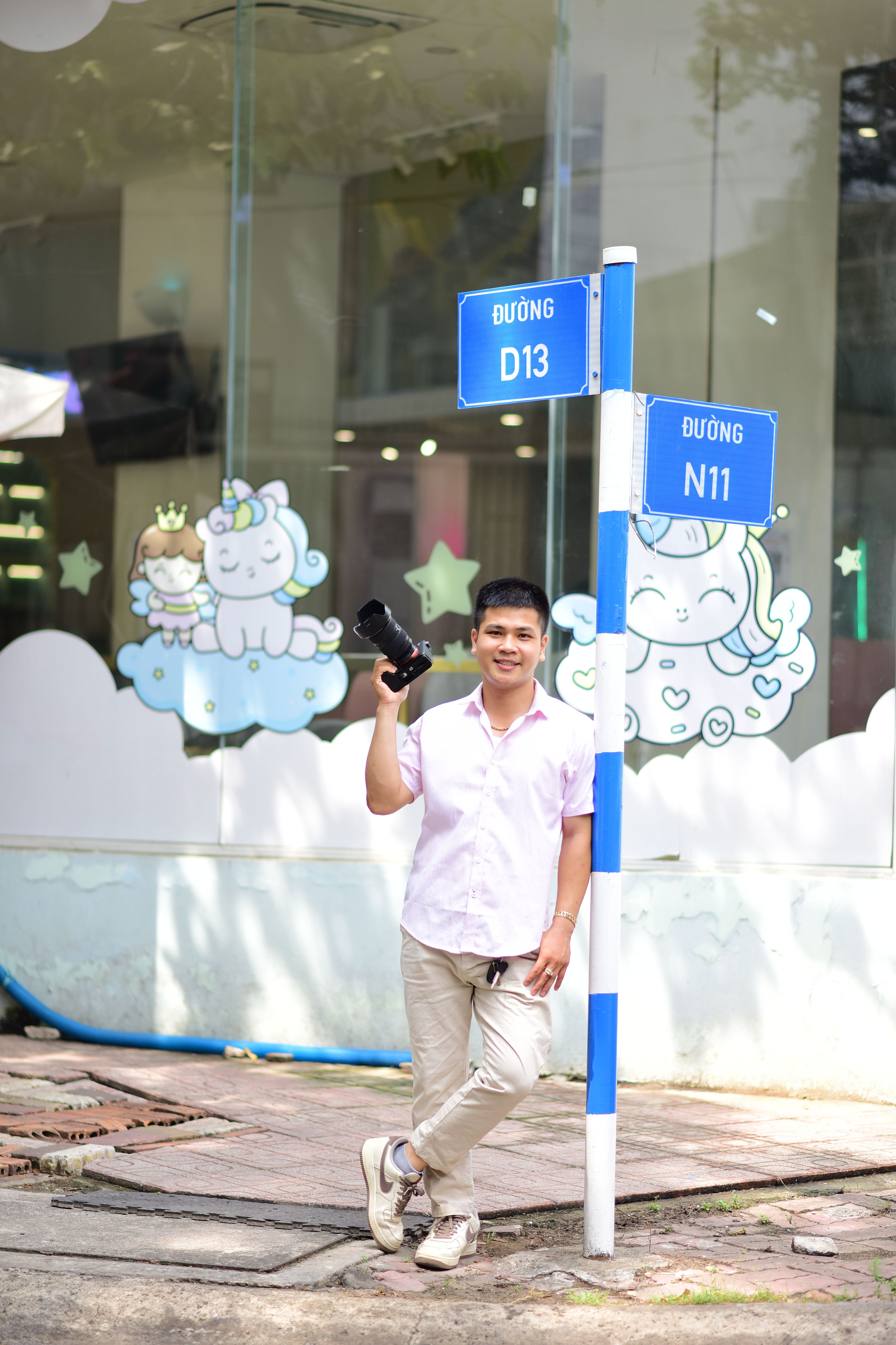A smiling man with a camera stands by a street sign with whimsical window art.