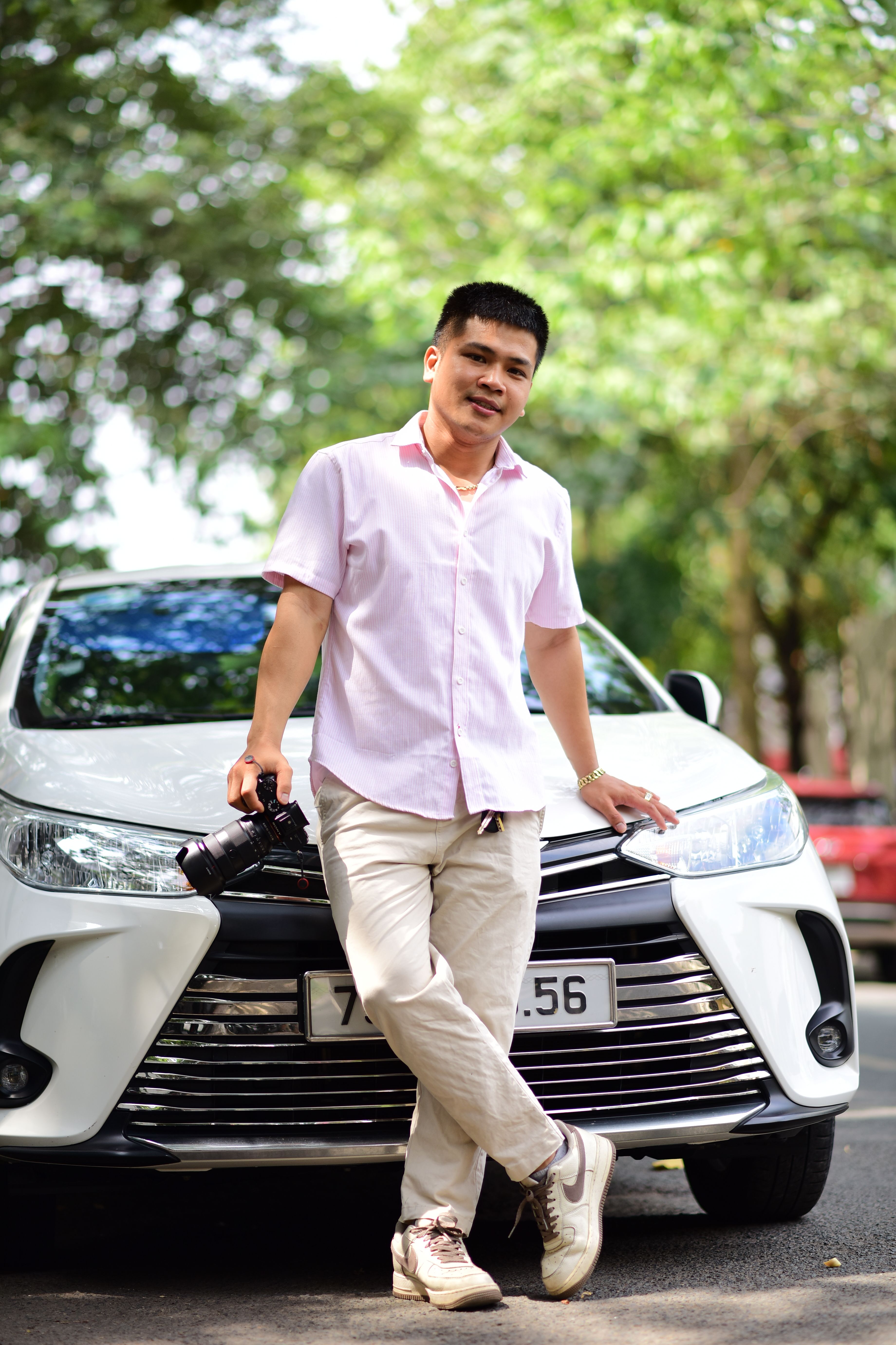 Man in a pink shirt leaning on a car outdoors with a smile.