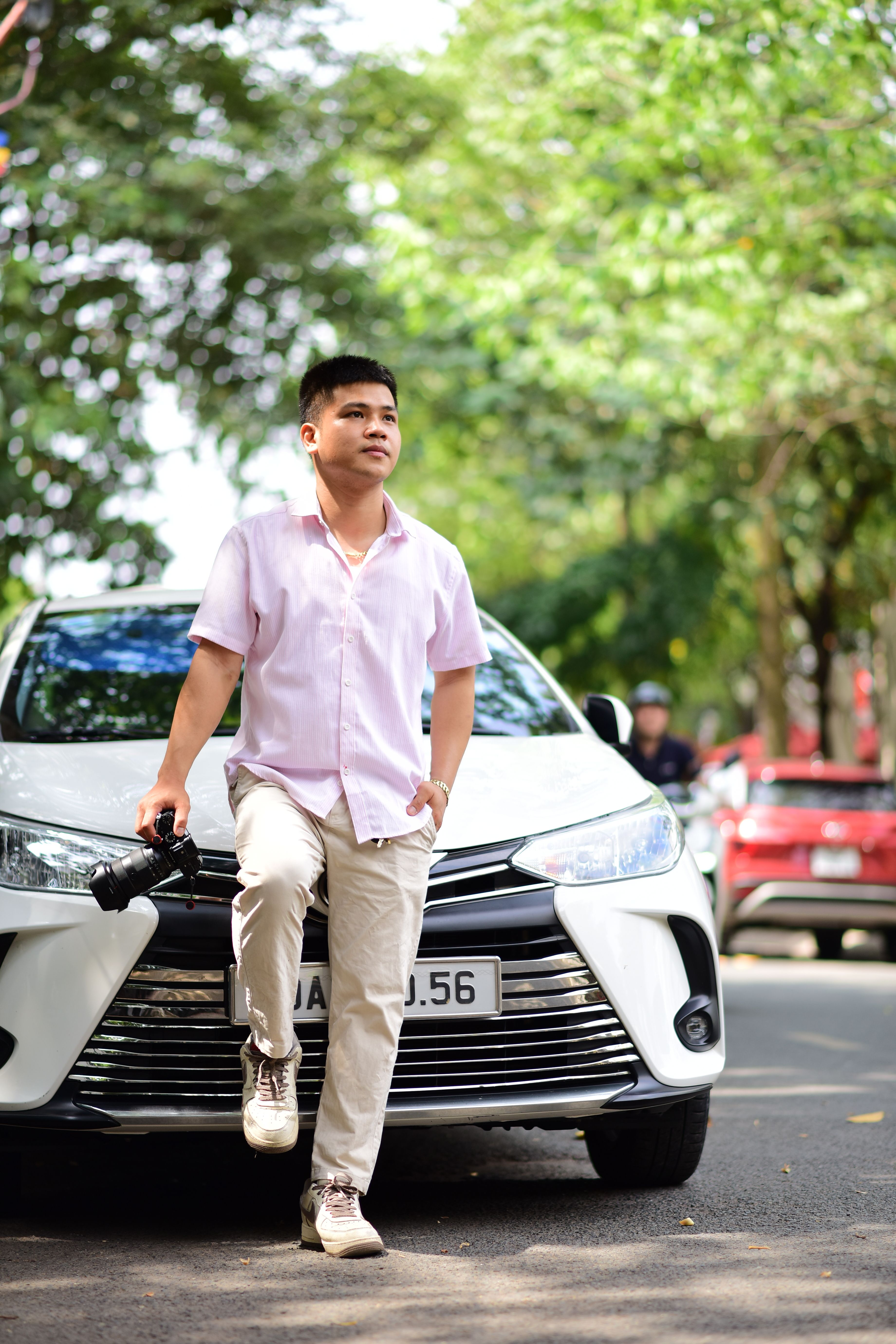 A man in casual attire leans on a white car with a camera, outdoors on a sunny day.