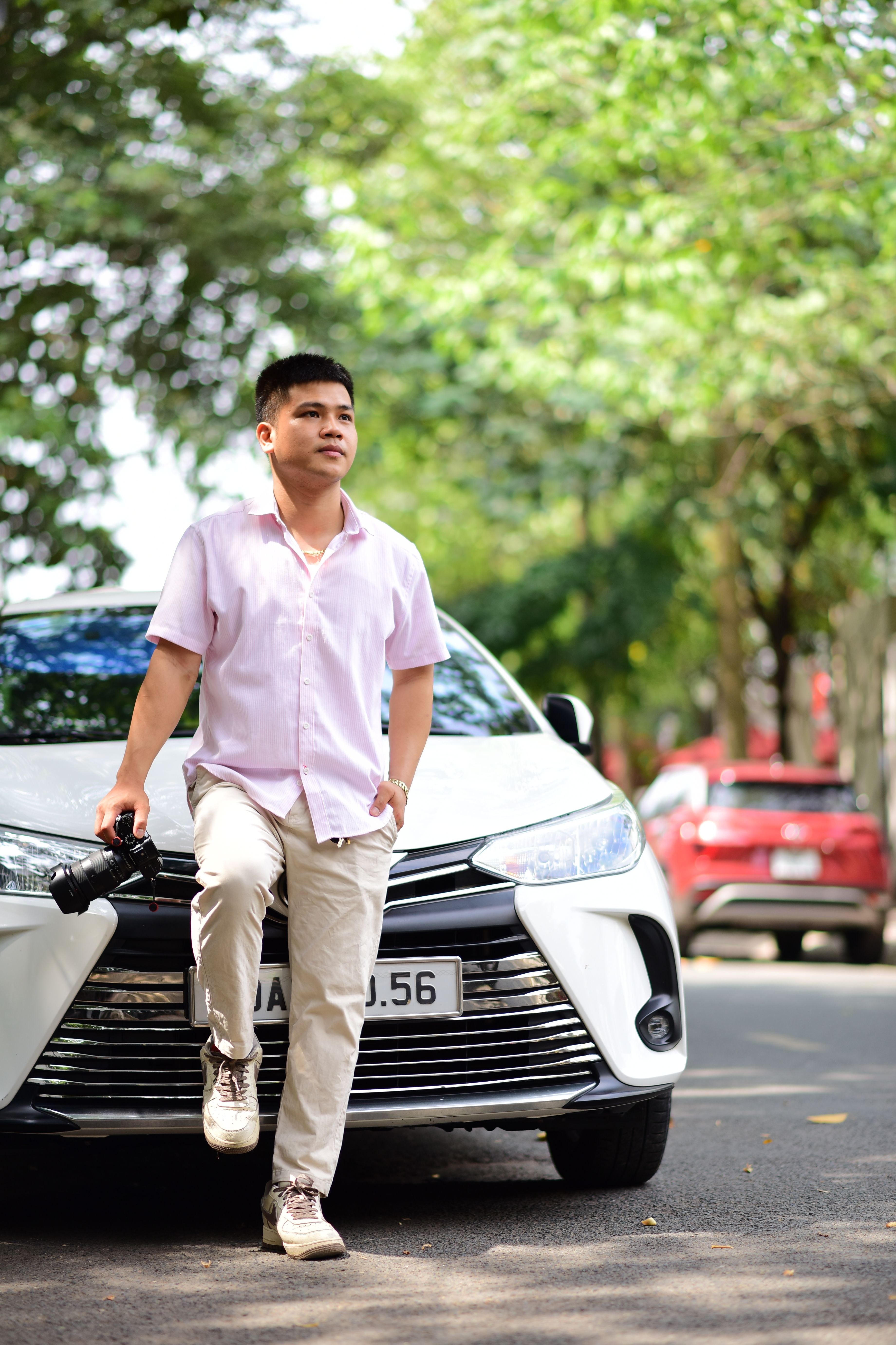 Casually dressed man with a camera poses by a car on a sunny street, exuding confidence and style.