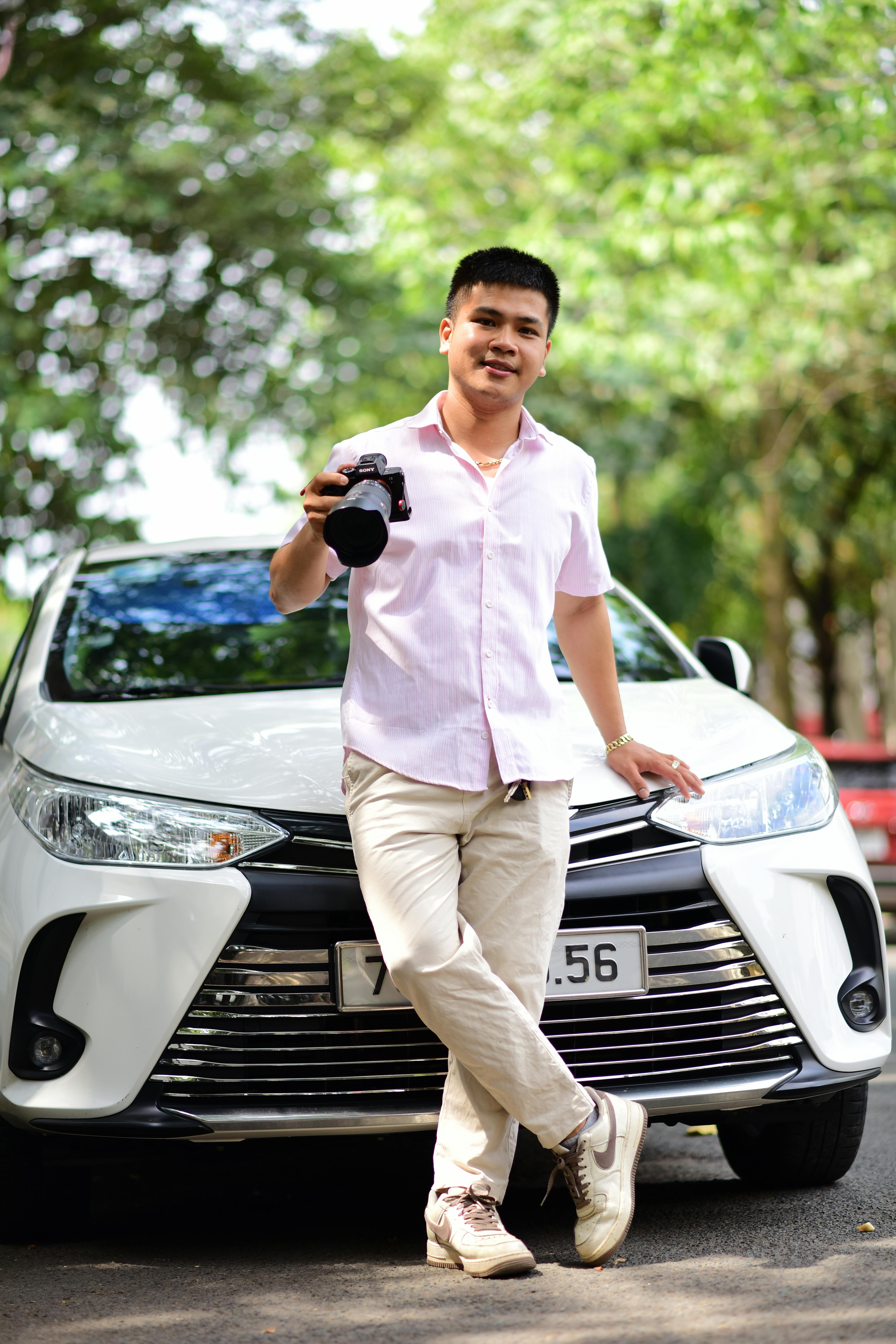 Young photographer holding camera near white car in a lush outdoor setting.