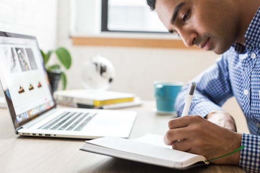Man focused on writing in a notebook at an office desk with a laptop and coffee cup.