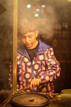 A young vendor roasts chestnuts on a bustling street in Istanbul at night, amidst the warm glow and smoke.
