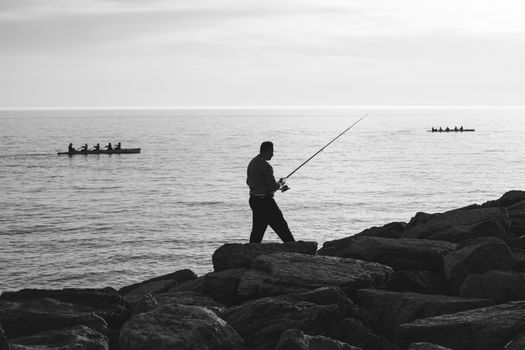 Man fishing on rocky shore with rowboats at sea during sunset. Serene coastal scene.