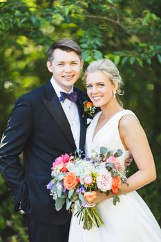 Beautiful outdoor wedding photo with smiling bride and groom in formal attire holding a colorful bouquet.