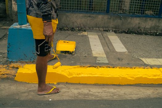 Street scene capturing a person in flip-flops painting a curb yellow with a roller.