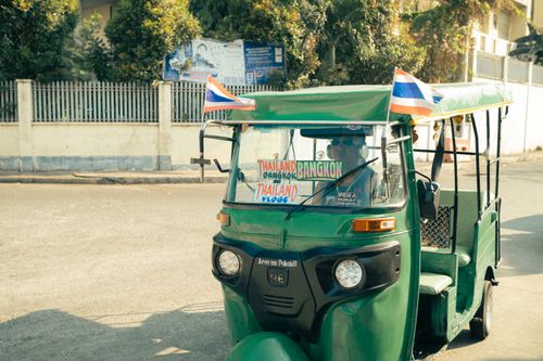 Free A traditional green tuk-tuk with a driver on a sunny street in Bangkok, Thailand, featuring Thai flags. Stock Photo