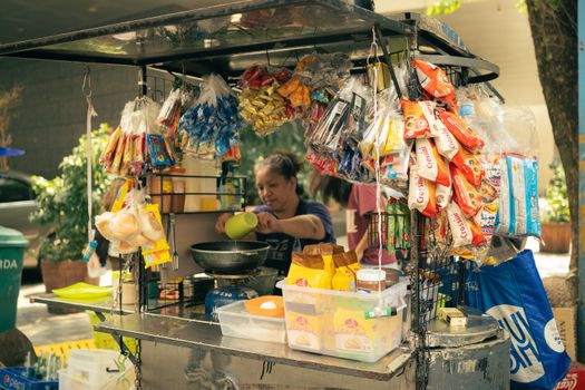 Street vendor preparing food among hanging snacks and colorful packaging outdoors during daytime.