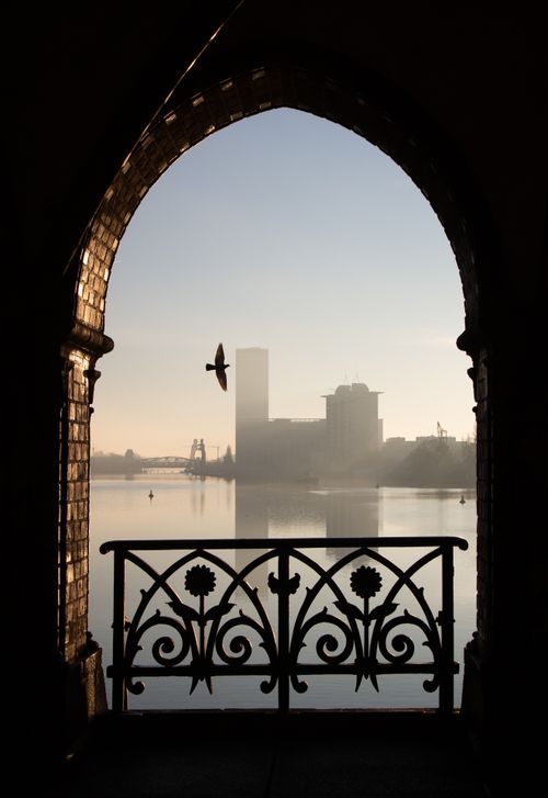 Free A tranquil view through an ornate archway highlights a foggy city skyline with a bird in flight. Stock Photo