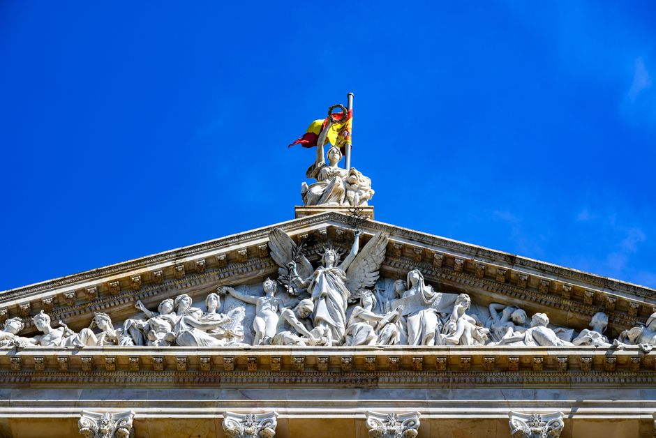 Arquitectura histórica de estilo partenon decorada con esculturas y bandera española, capturada en vibrante luz solar.