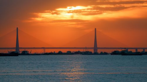 Free Silhouetted ships and bridge under a vibrant orange sunset in an industrial harbor. Stock Photo