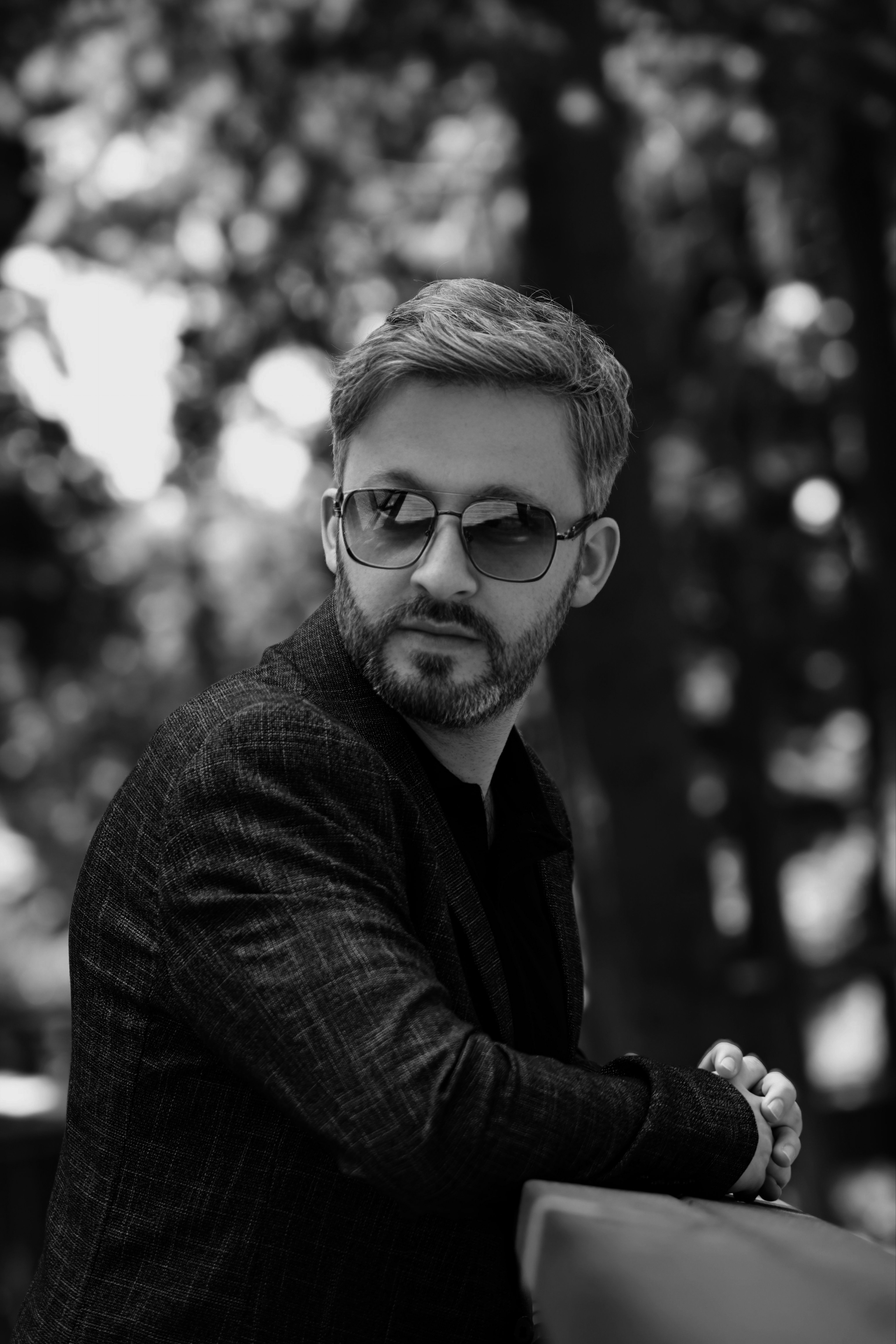 Black and white portrait of a stylish man wearing sunglasses and leaning on a railing outdoors.