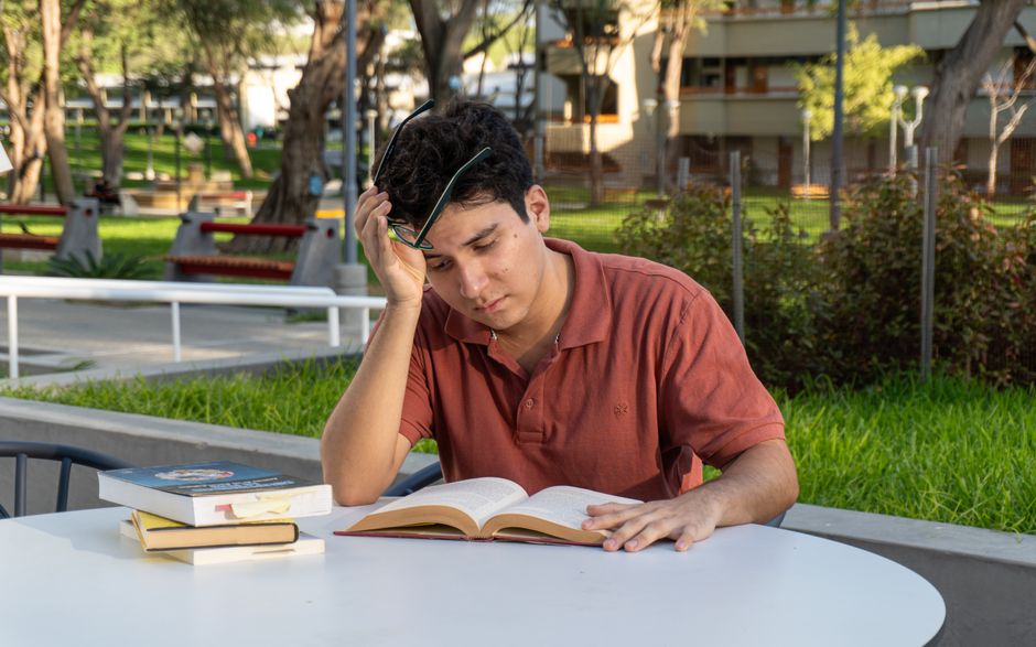 A young man is studying outdoors in a park, focused on reading a book at a table. A young man is studying outdoors in a park, focused on reading a book at a table.