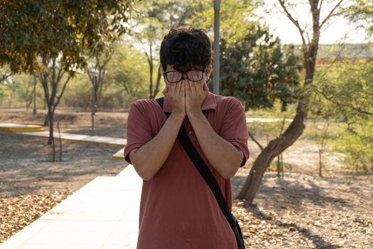 A young man stands in an outdoor park with trees covering his face with his hands.