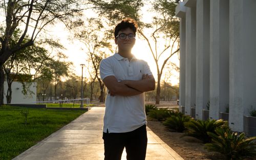 Free A young man stands confidently on a university campus at sunset, framed by modern architecture and lush trees. Stock Photo