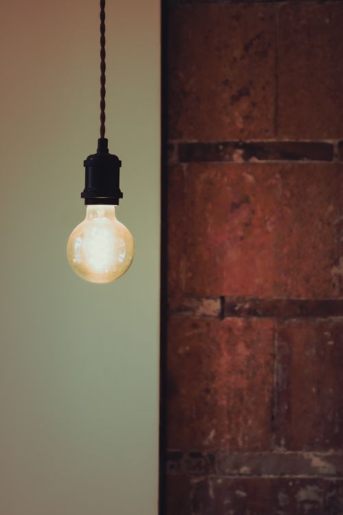 Free A glowing vintage light bulb hanging against a rustic brick wall, creating a warm ambience. Stock Photo