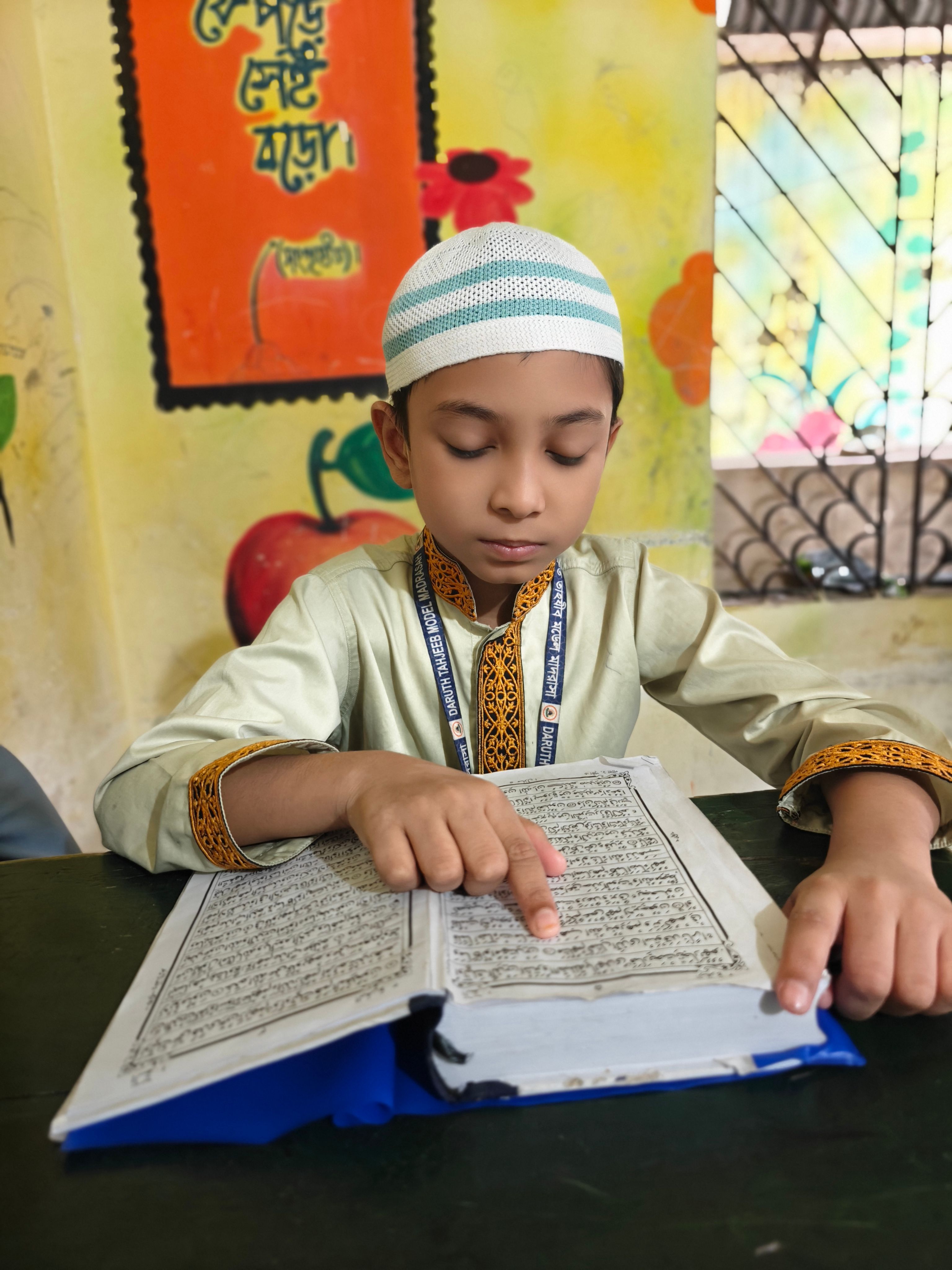A young boy in traditional attire reading a religious book inside a classroom.