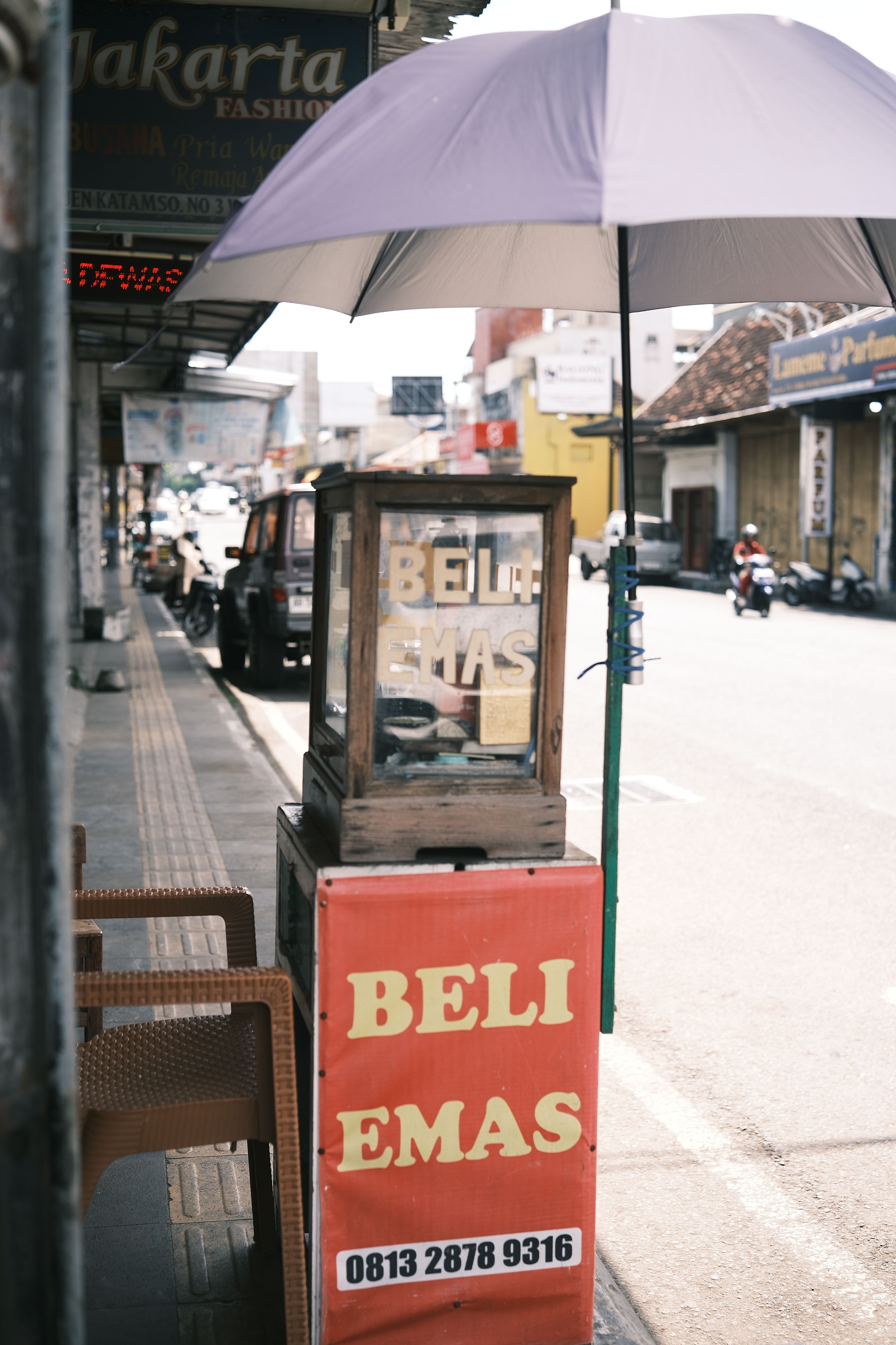 Fotomural Escena urbana de un puesto de venta ambulante con un cartel de 'beli emas' bajo una sombrilla.