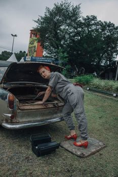 A woman in gray attire repairs a vintage car's trunk in an outdoor setting, showcasing DIY skills.