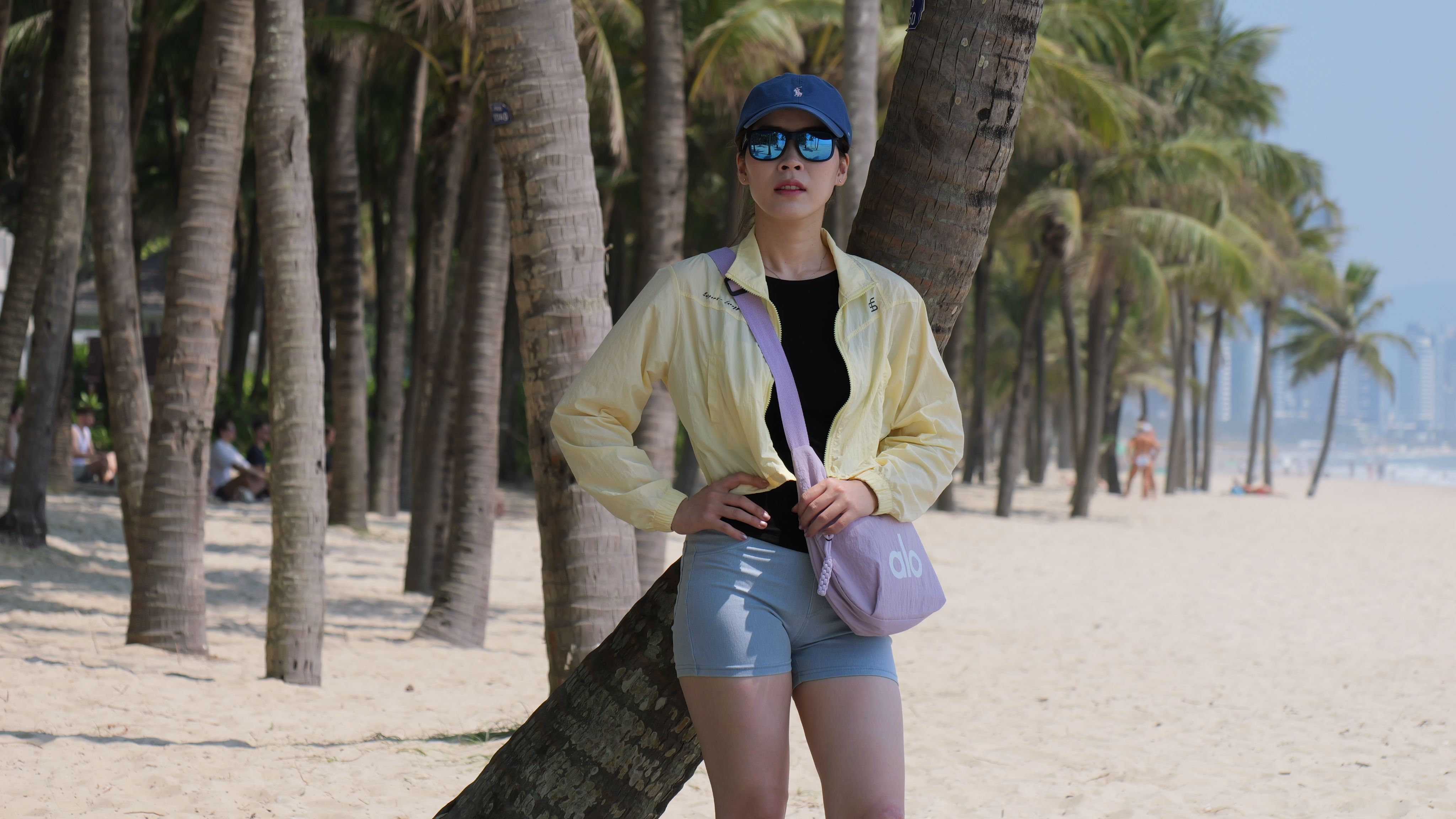 Woman in casual wear leaning against palm tree on a sunny tropical beach, wearing a hat and sunglasses.