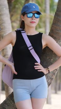 Woman in black top, blue shorts, and cap standing by a palm tree, exuding a relaxed vibe.