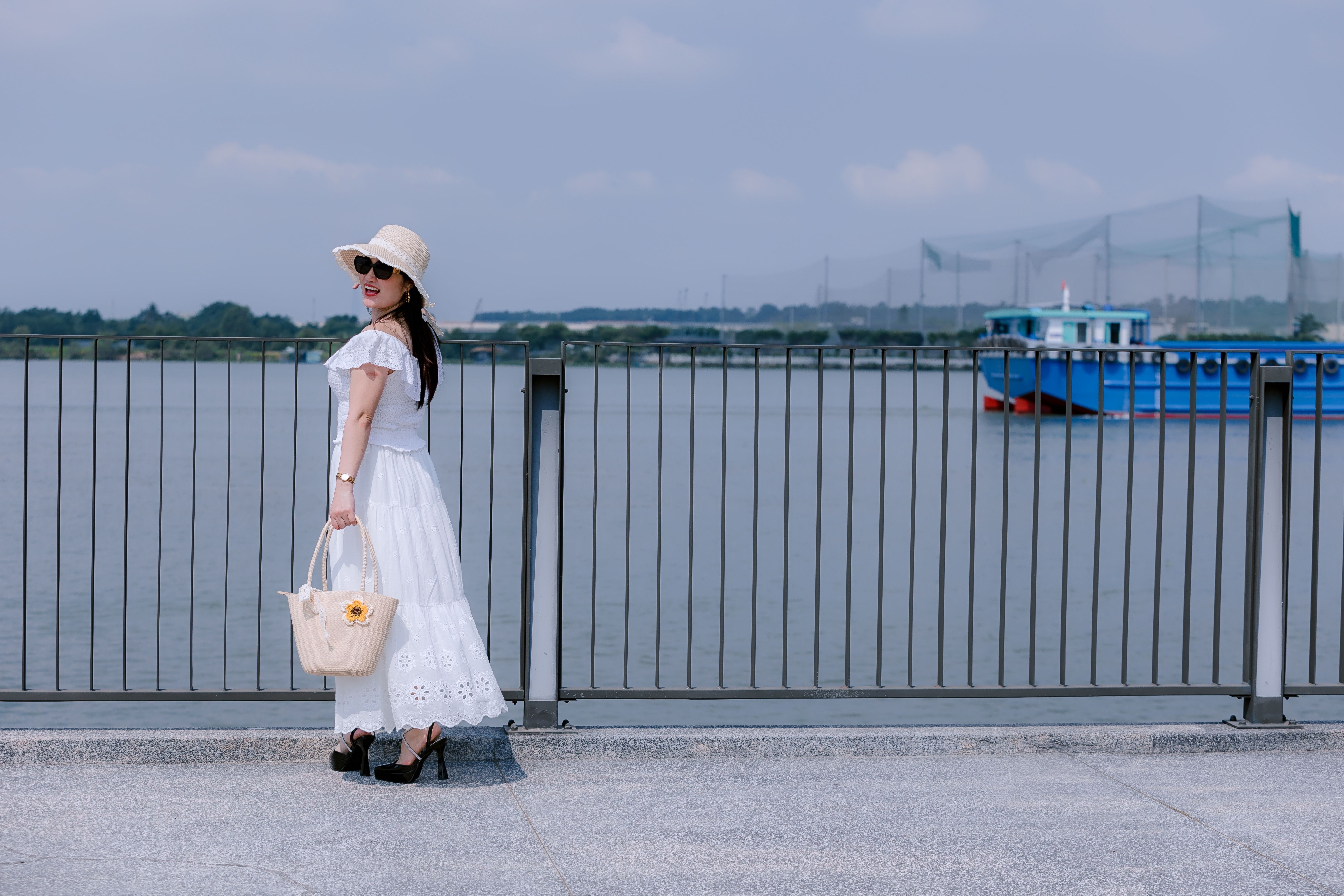 Fashionable woman in white dress by a waterfront with a blue boat passing by.