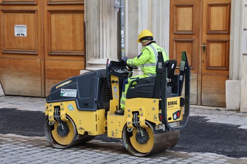 Free A construction worker in safety gear operates a road roller on a cobblestone street near a historical building. Stock Photo
