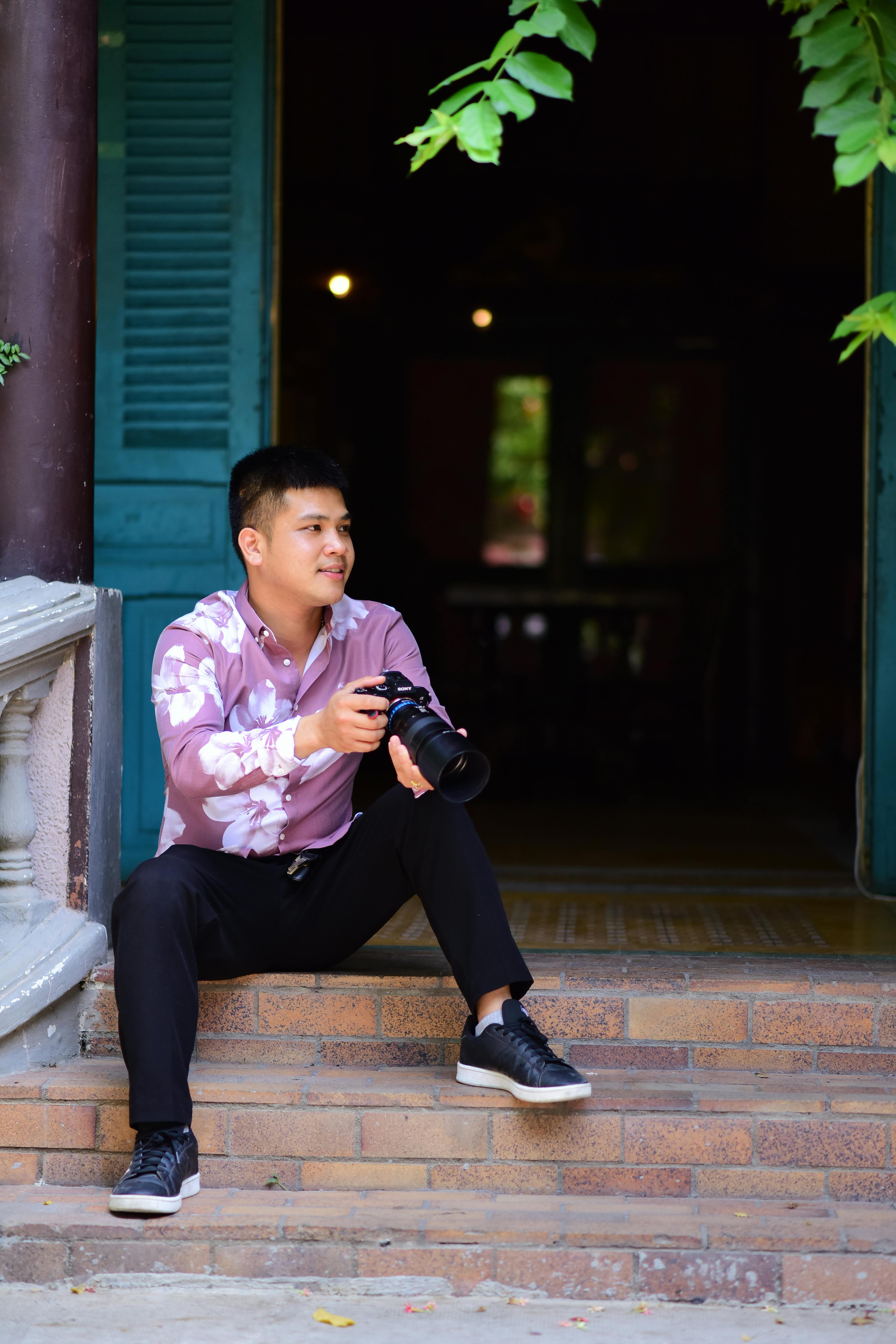 Young photographer sitting on steps with a camera, smiling, in an indoor setting with natural light.