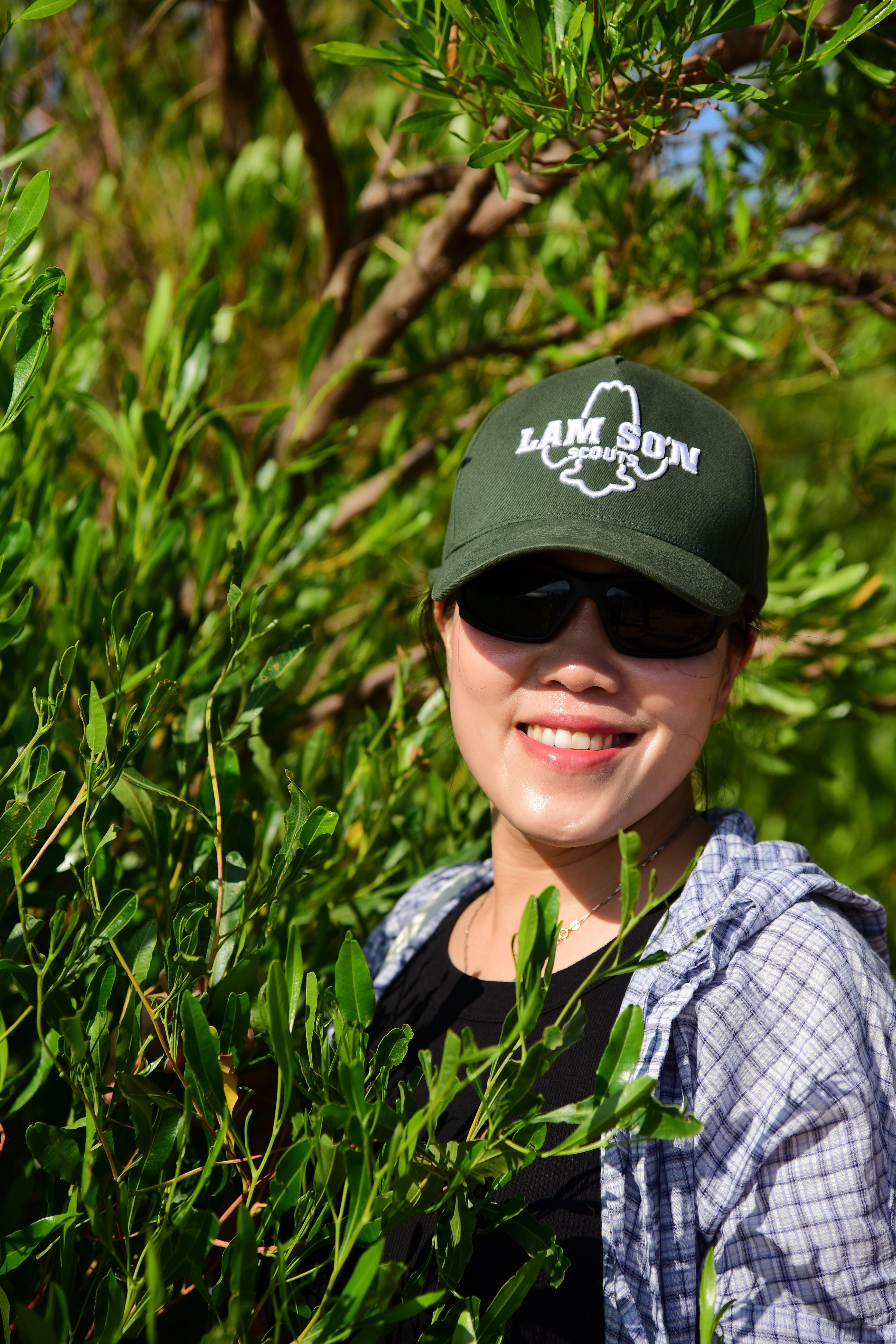 Woman wearing sunglasses and cap surrounded by green foliage on a sunny day.