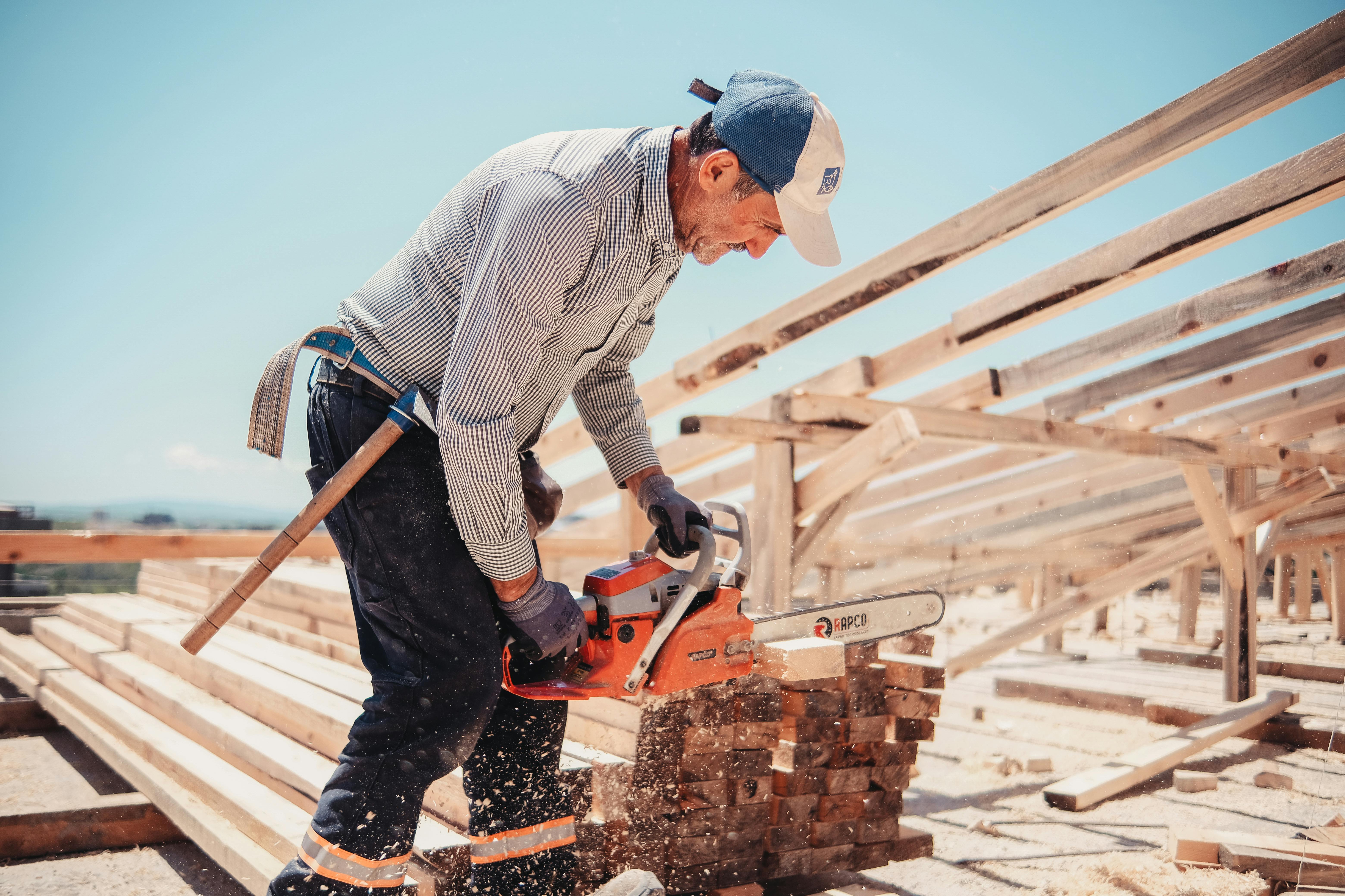 Charpentier qualifié utilisant une tronçonneuse pour couper du bois sur un chantier de construction en extérieur, sous un ciel dégagé.
