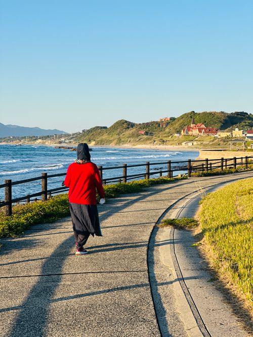 Free A woman in red walks along a coastal path with scenic seaside views during sunset. Stock Photo