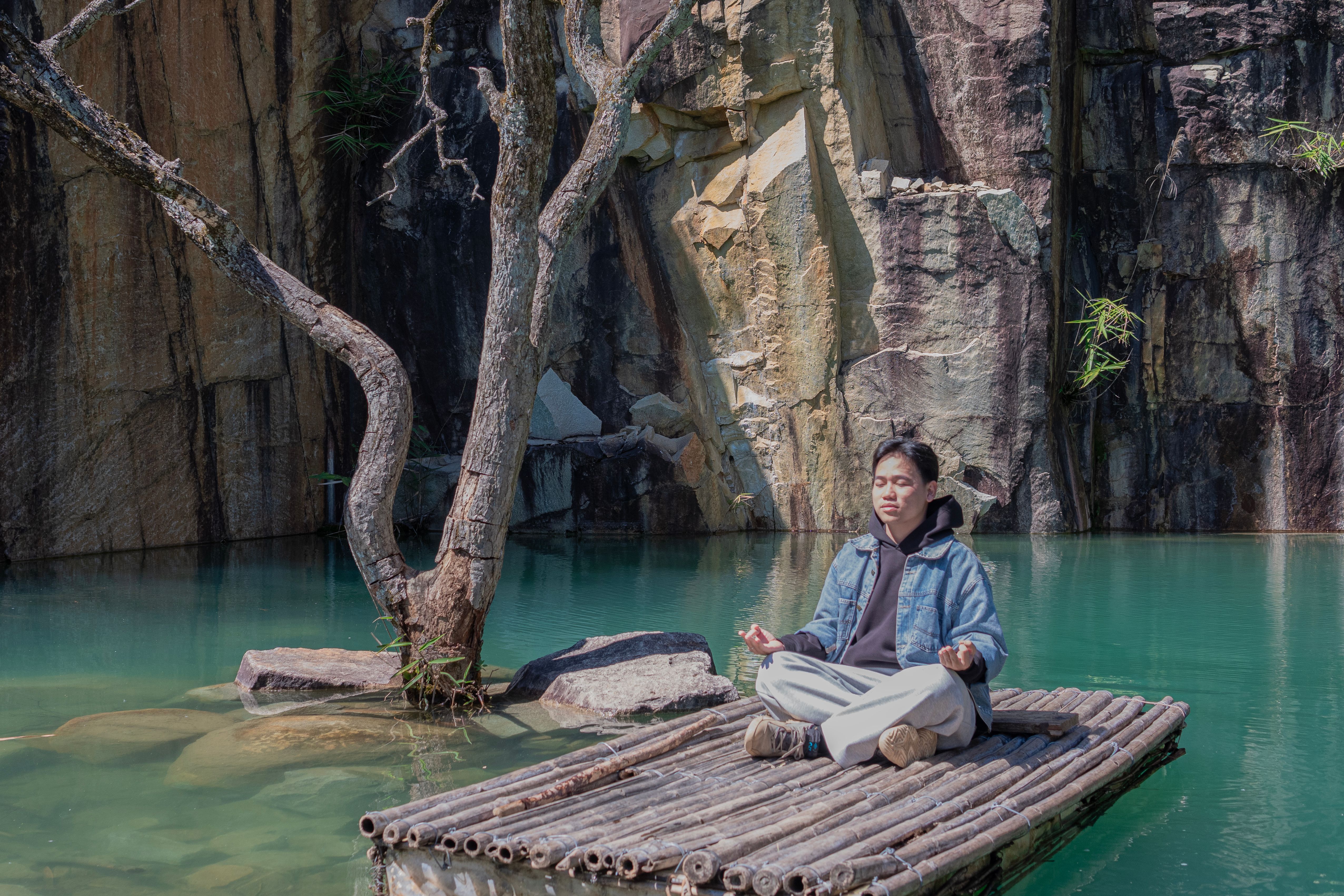 A serene scene of a man meditating on a bamboo raft by a scenic cliffside water body.