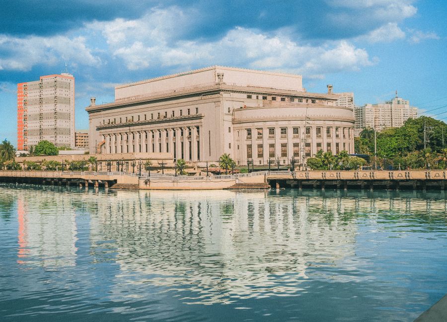 The iconic Manila Post Office building reflecting on the Pasig River under a bright blue sky.