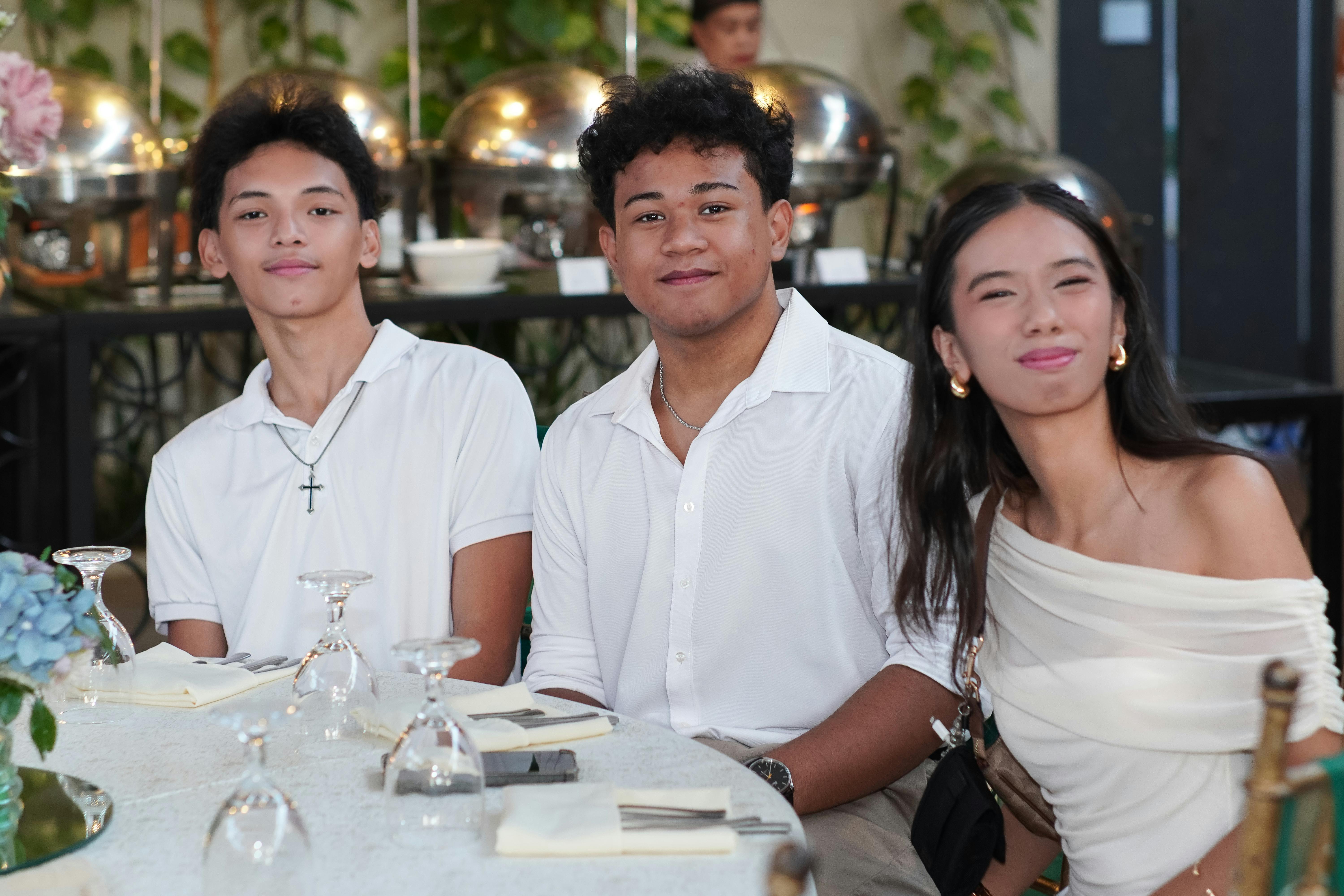 Three young adults smiling at a table during a formal gathering indoors, captured in a lively setting.