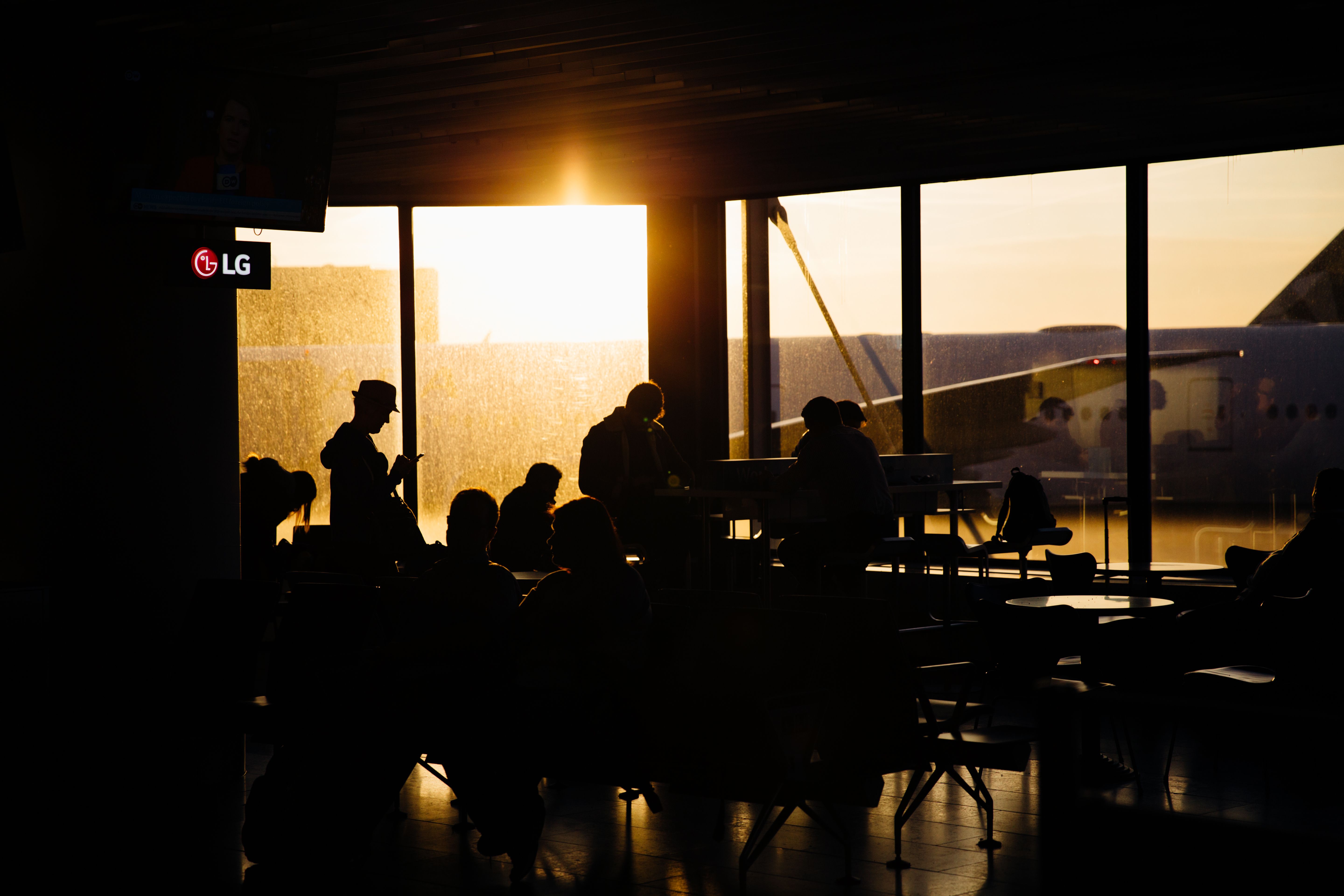 Passengers wait at an airport departure gate as the setting sun shines through the terminal windows.