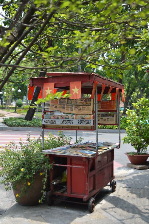 Free Colorful newspaper stand with star flags in a leafy park setting, evoking local culture. Stock Photo