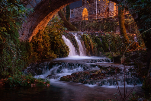 Free A tranquil waterfall flows under an ancient stone bridge, surrounded by lush greenery in Λιβαδειά, Ελλάδα. Stock Photo