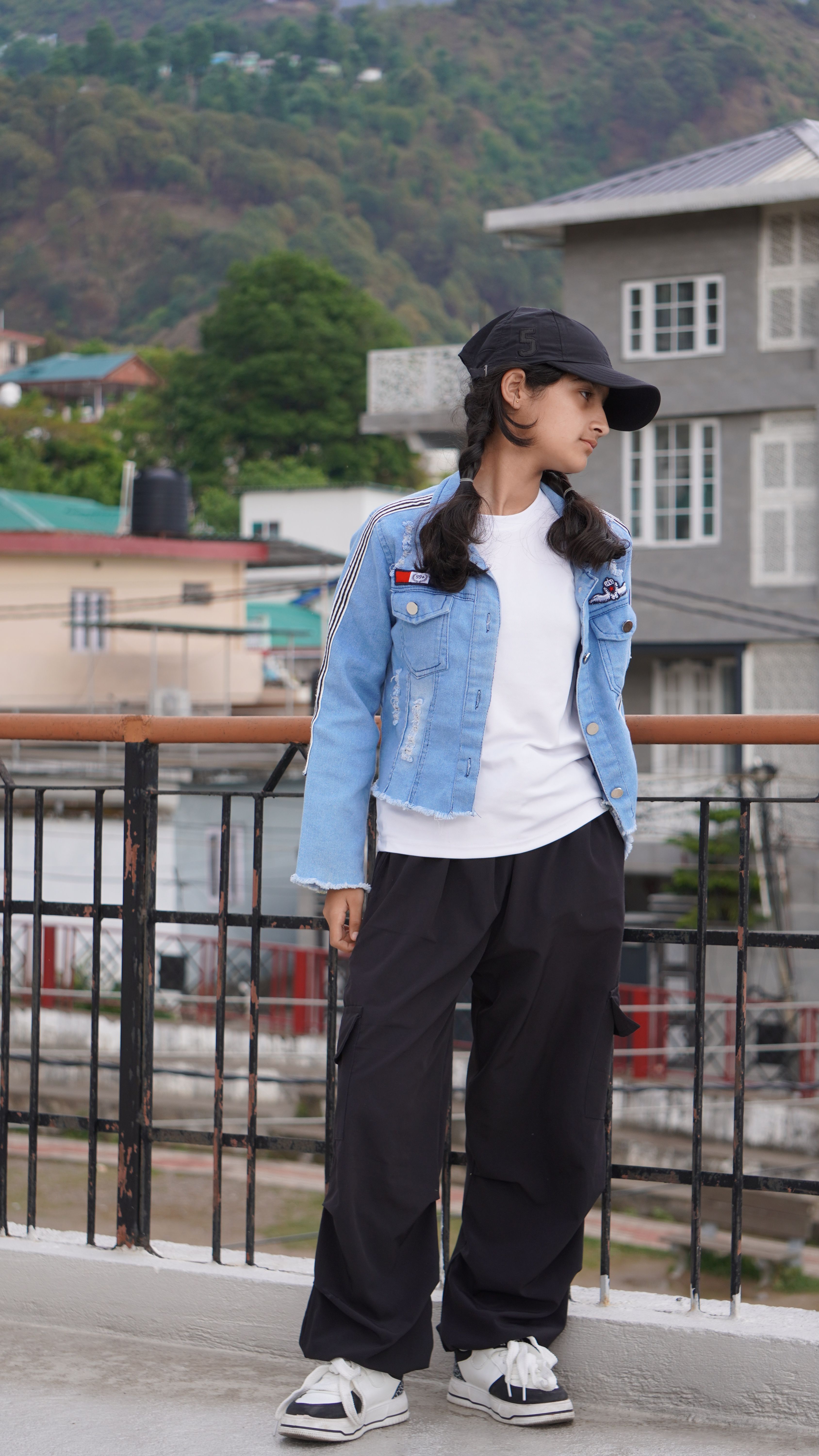 Teenager in modern fashion poses outdoors in Dharamshala, showcasing urban style with mountains in the background.