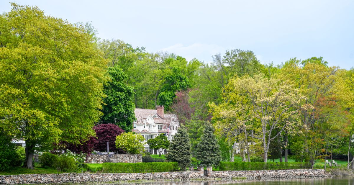Idyllic countryside house surrounded by lush trees reflected in a calm lake during springtime.