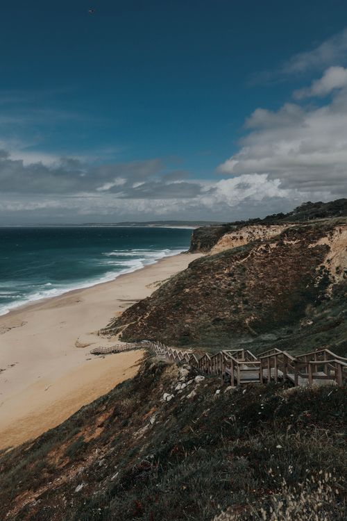 Free Scenic view of a coastal beach with rugged cliffs and a wooden walkway under dramatic skies. Stock Photo