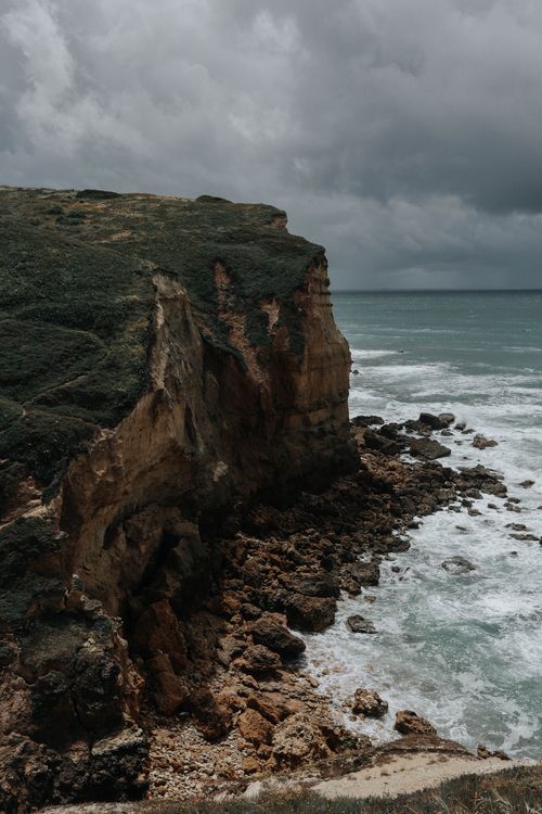 Free A steep coastal cliff with rocky shore beneath a dramatic, cloudy sky, highlighting nature's rugged beauty. Stock Photo