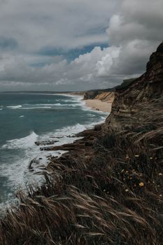 Dramatic view of a coastal cliff with rugged rocks and ocean waves under cloudy skies.